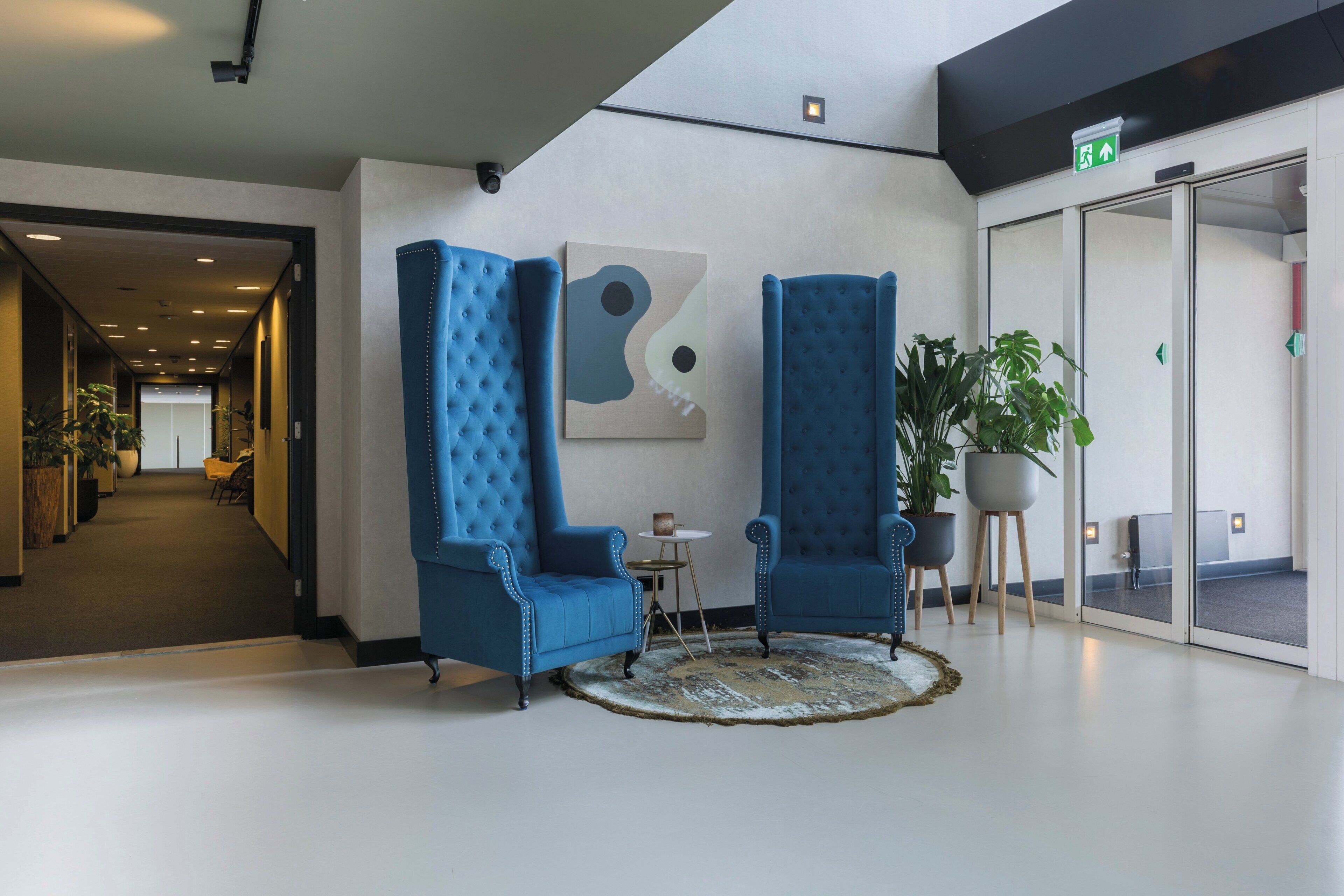 A lobby with two blue armchairs, a round rug, a painting, and a potted plant.