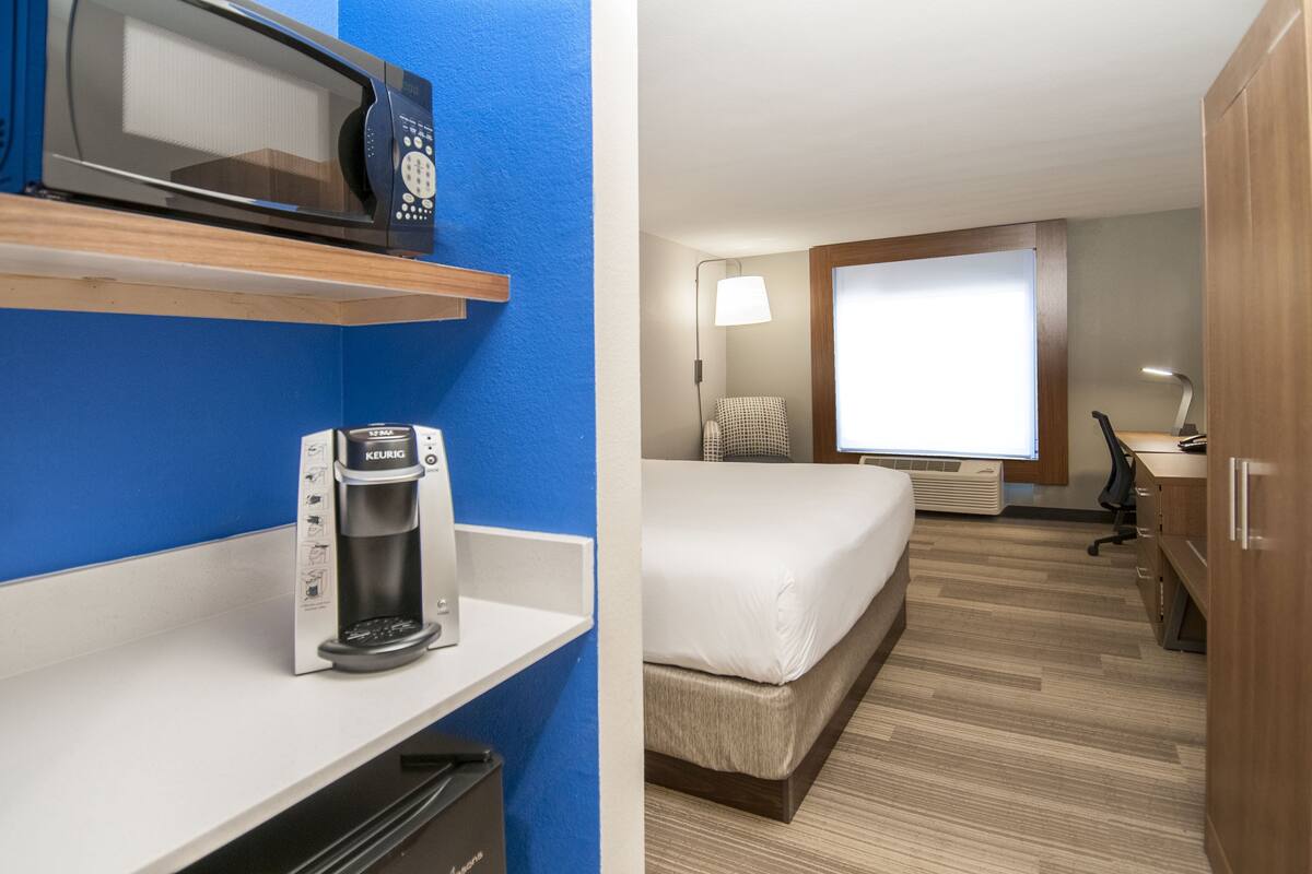 A hotel room with a microwave, coffee maker, bed, desk, lamp, and window.