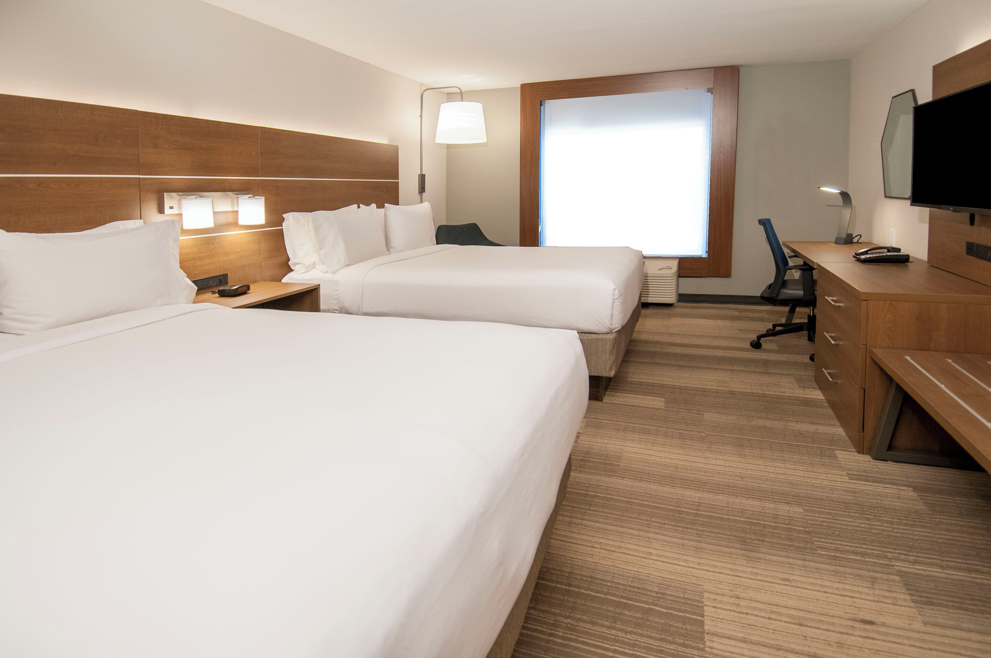 Quarto de hotel com duas camas, uma escrivaninha, uma cadeira, uma televisão e uma janela com persianas.