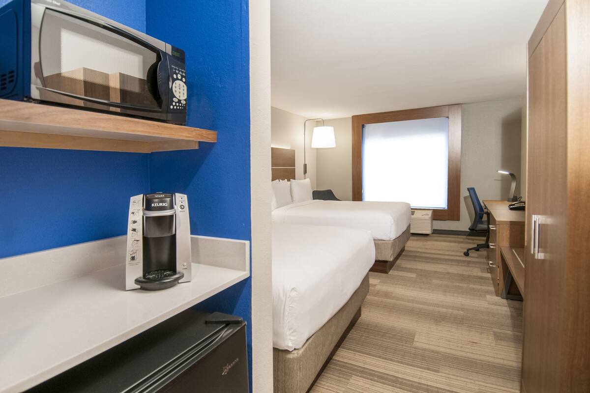 A hotel room with a microwave, coffee maker, two beds, a desk, and a window.