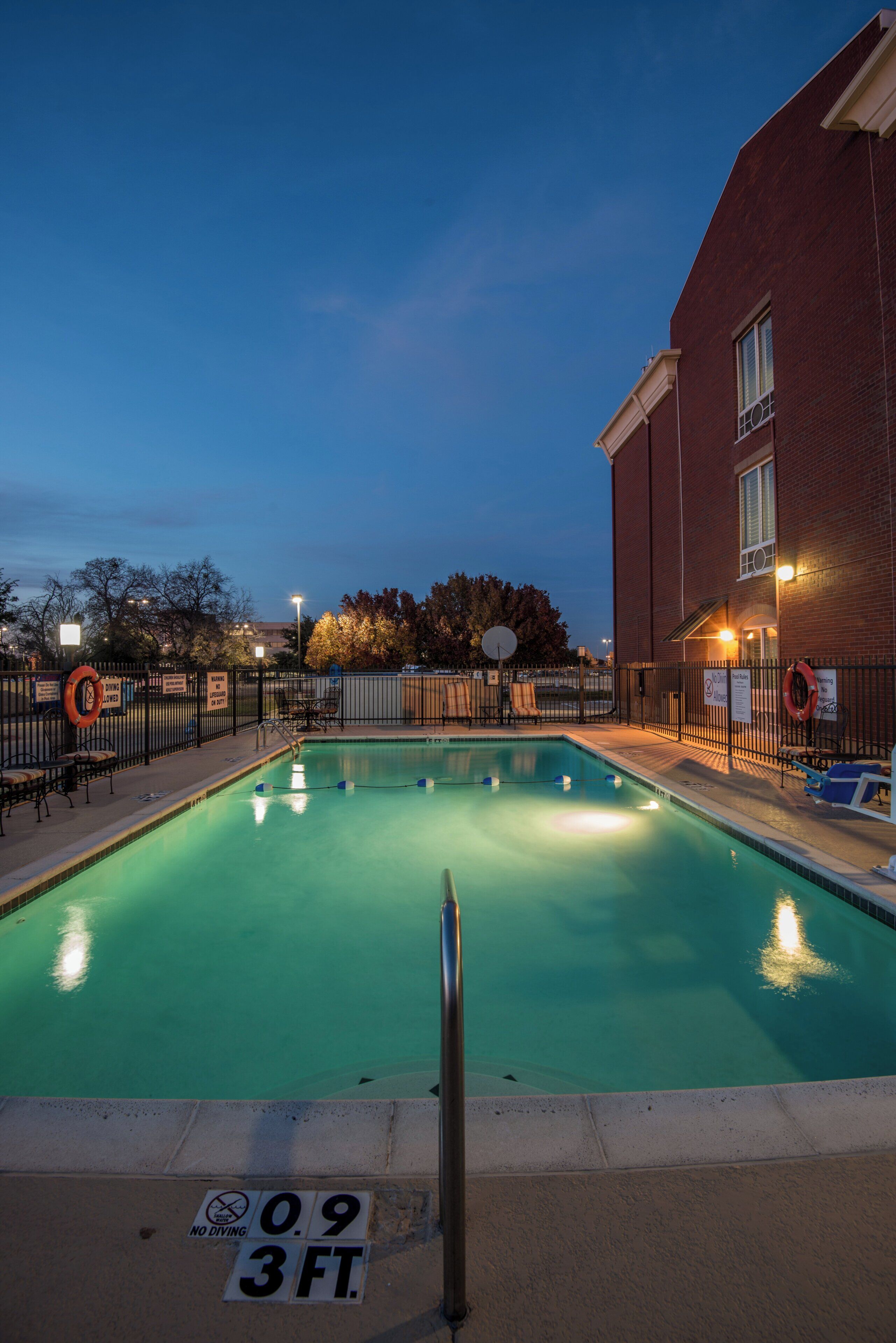 A swimming pool with a 3 FT depth sign, surrounded by a fence, and adjacent to a brick building.