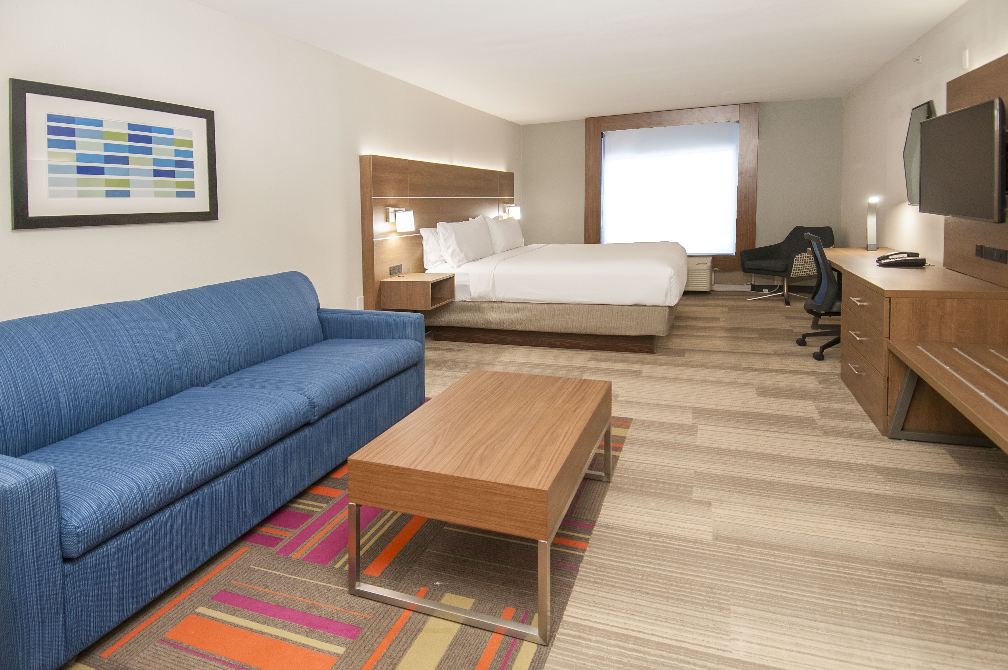 A hotel room with a bed, a blue sofa, a wooden desk, a television, and a colorful rug.