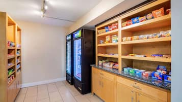 A kitchenette with a refrigerator, shelves stocked with snacks, and a countertop with packaged goods.