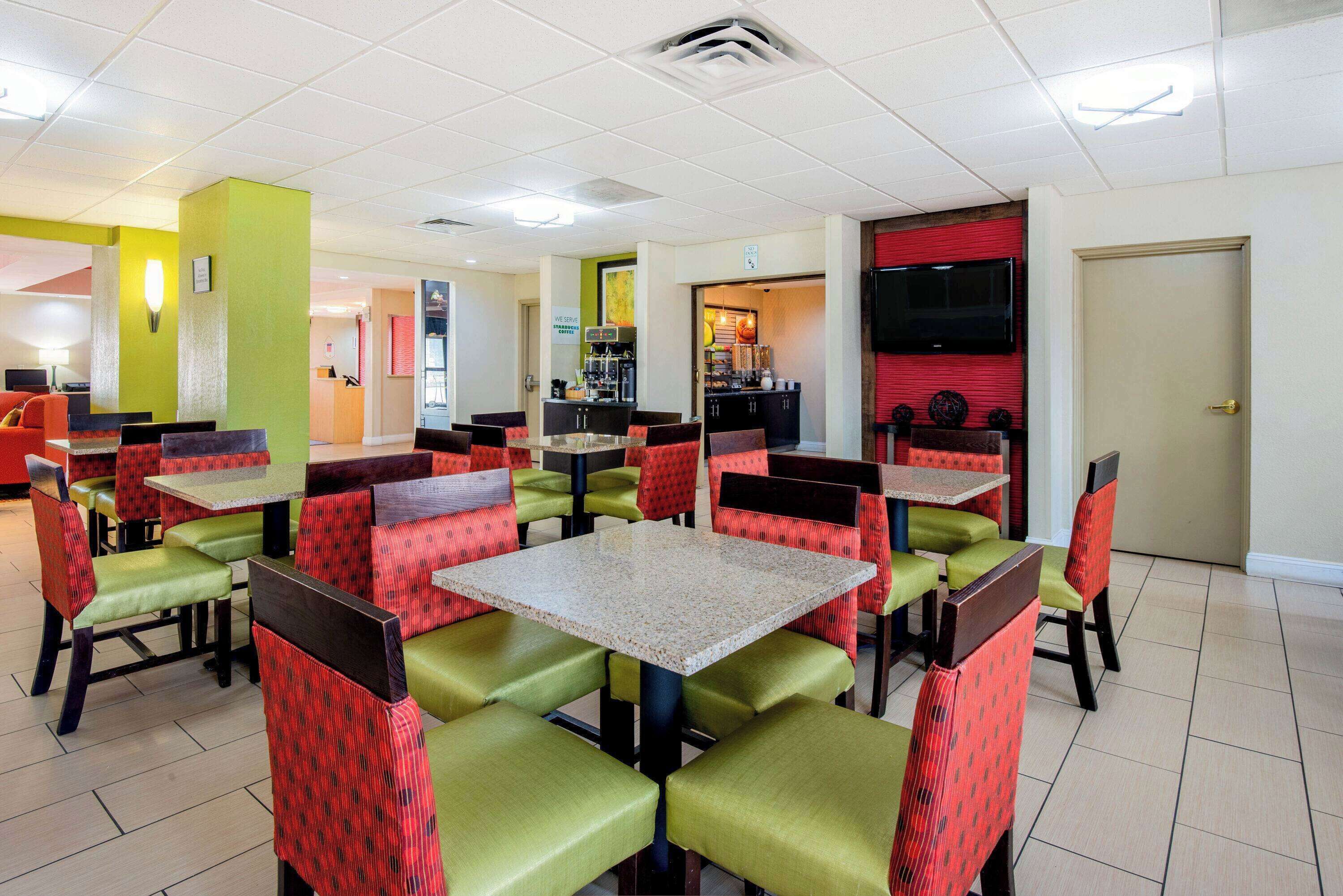 A restaurant with green and red upholstered booths and tables, a kitchen area in the background, and a door to the right.