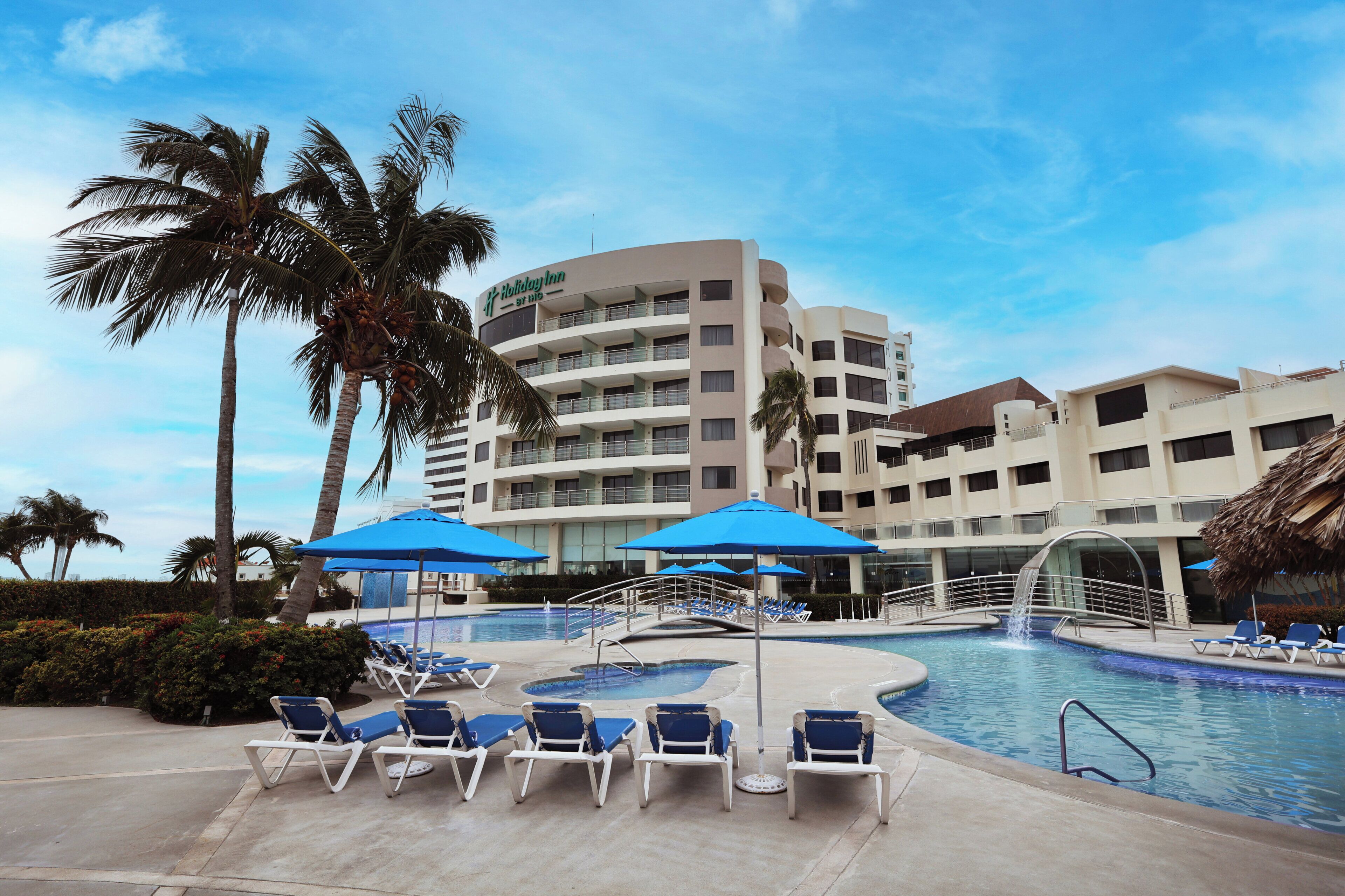 Un hotel con zona de piscina, sillas de descanso y sombrillas, palmeras y cielo despejado.