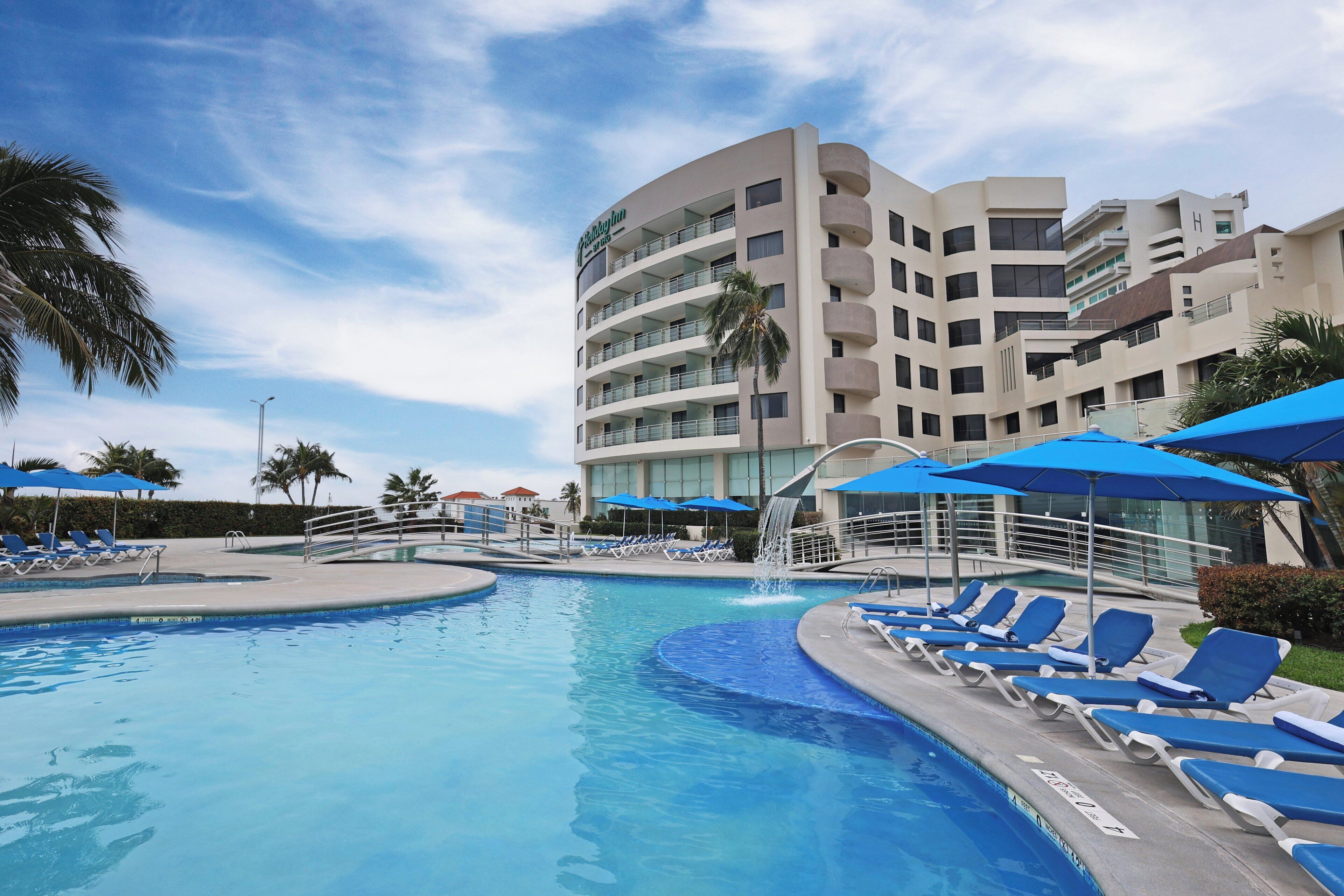 A hotel pool area with lounge chairs and umbrellas, a palm tree, and a modern building with balconies.