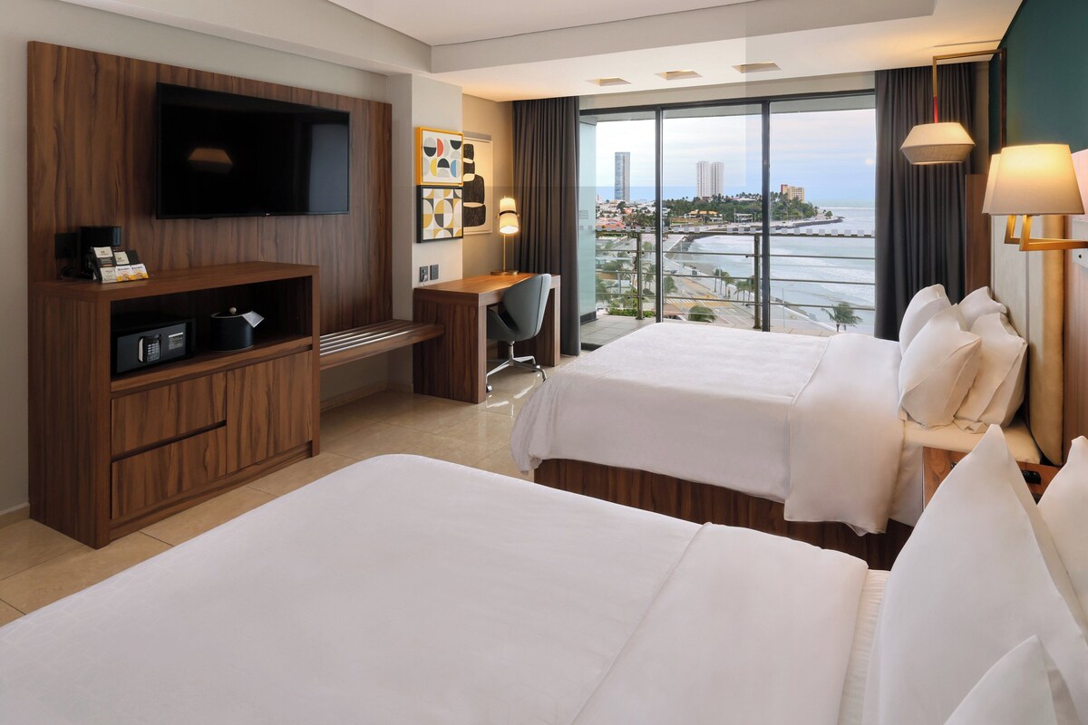 A hotel room with two beds, a desk, a TV, and a view of the city and sea.