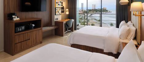 A hotel room with two beds, a desk, a TV, and a view of the city and sea.