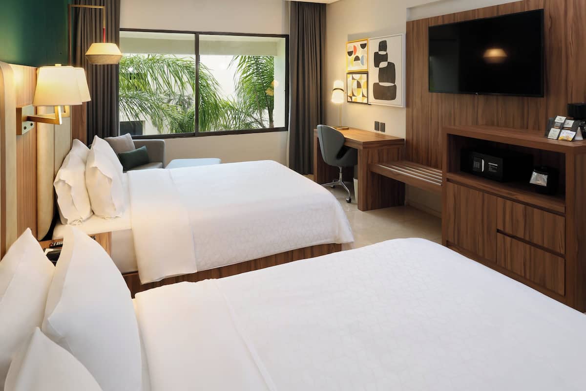 A hotel room with two beds, a desk, a chair, a TV, and a large window with curtains.