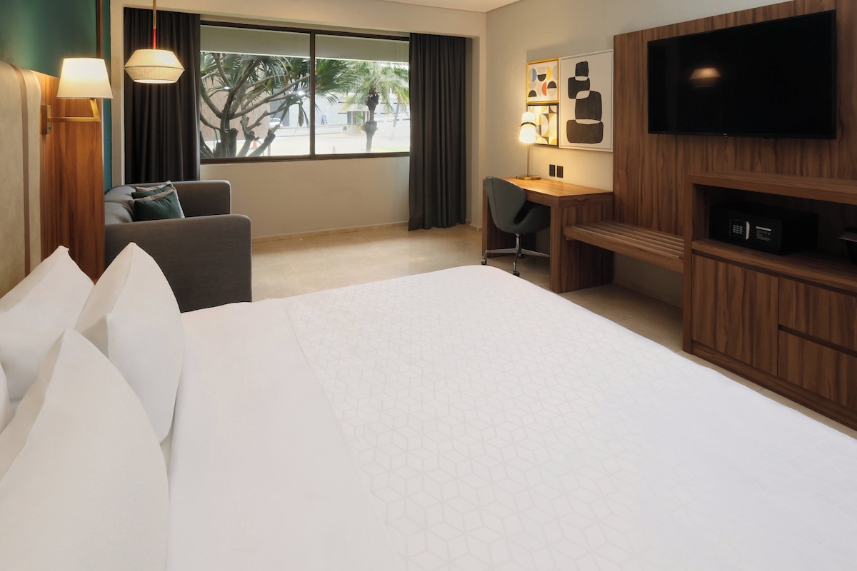 A hotel room with a large bed, a desk with a chair, a TV, and a window with curtains.