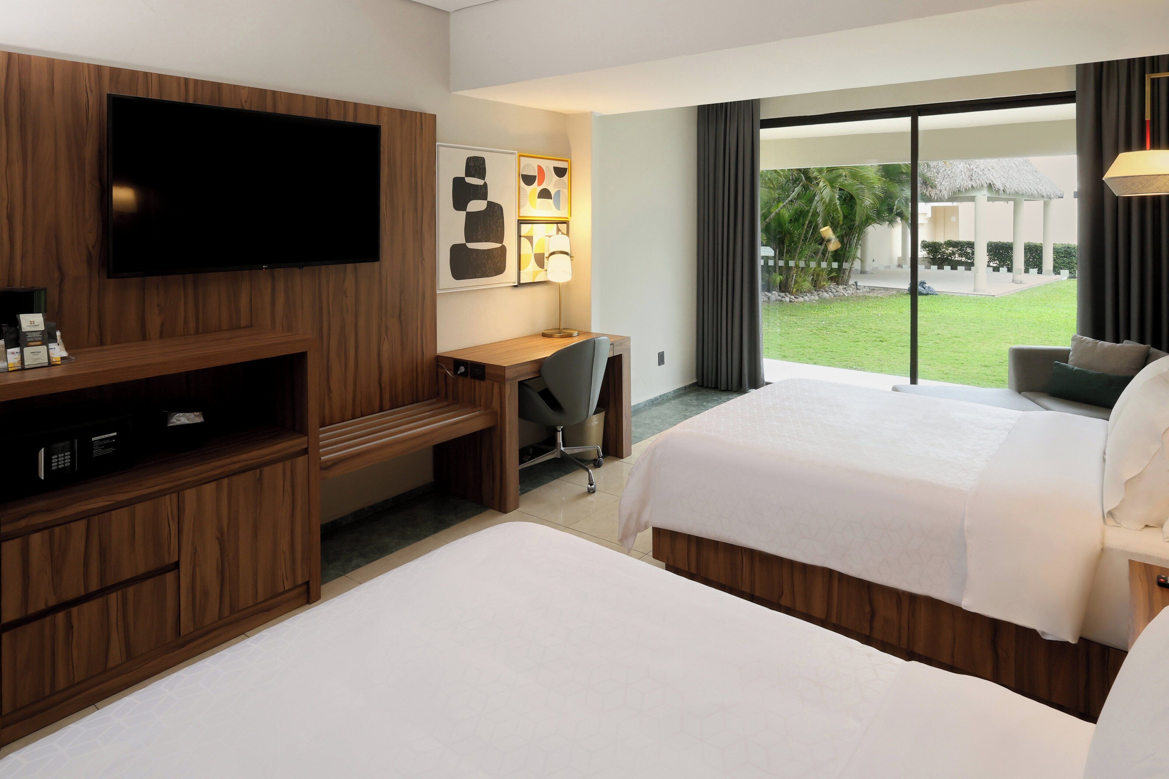 A hotel room with two beds, a desk, a TV, and a view of a garden.