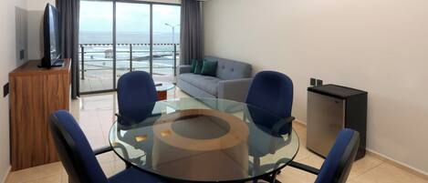 A modern hotel room with a glass-top dining table, blue chairs, a sofa, a TV, and a view of the ocean.
