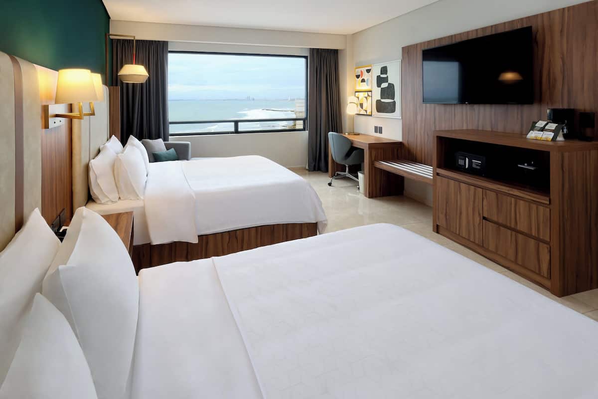 A hotel room with two beds, a desk, a TV, and a view of the ocean.
