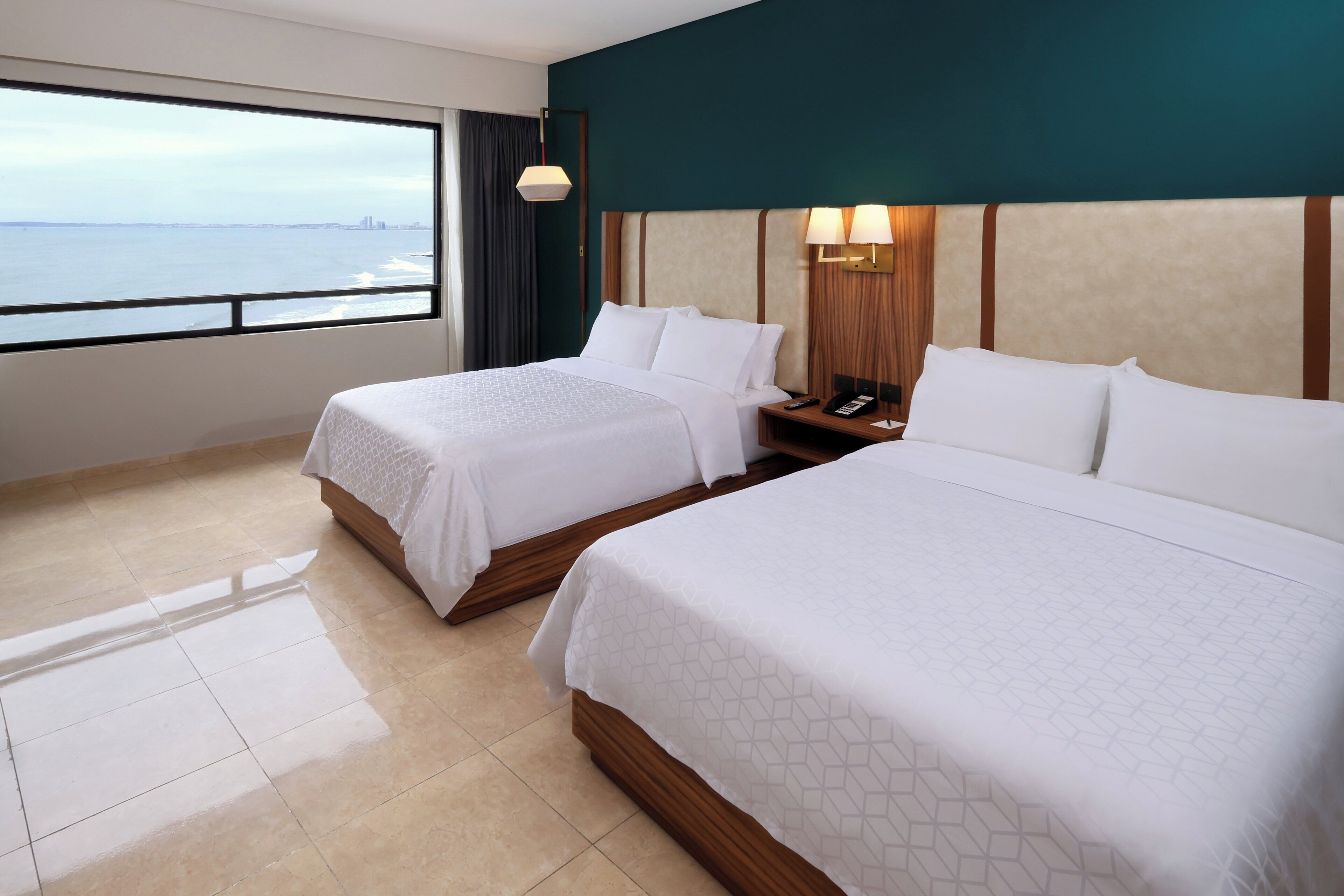 A hotel room with two beds, a large window with a view of the sea, and a nightstand with a lamp.