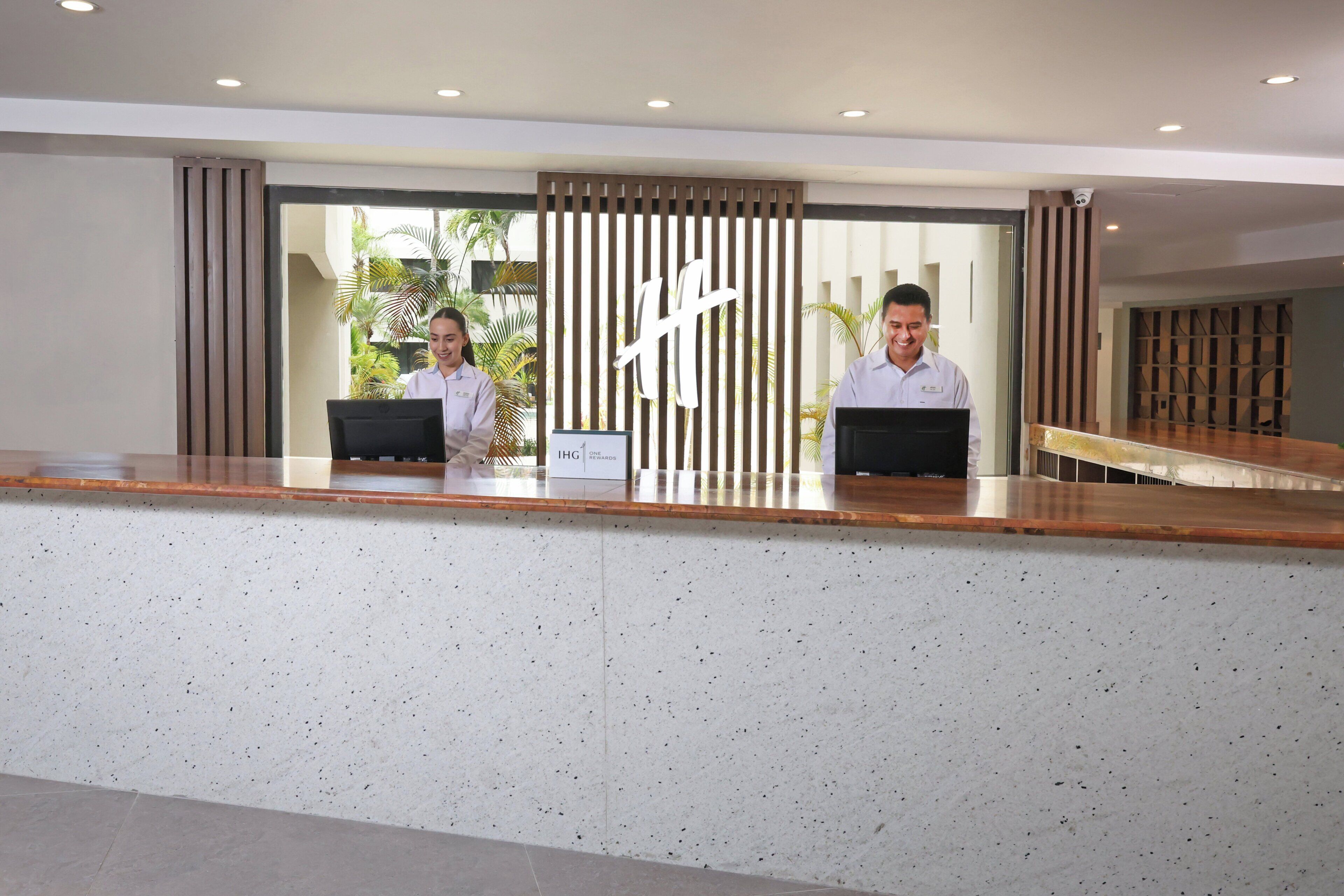 A reception desk with two staff members, a sign indicating 'IHG', and a view of the outdoors with plants.