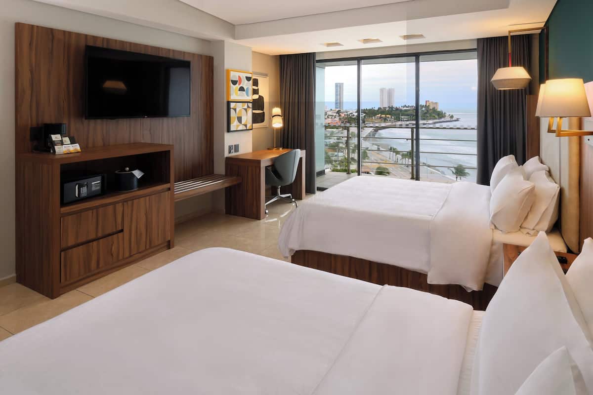 A hotel room with two beds, a desk, a TV, and a view of the city and sea.