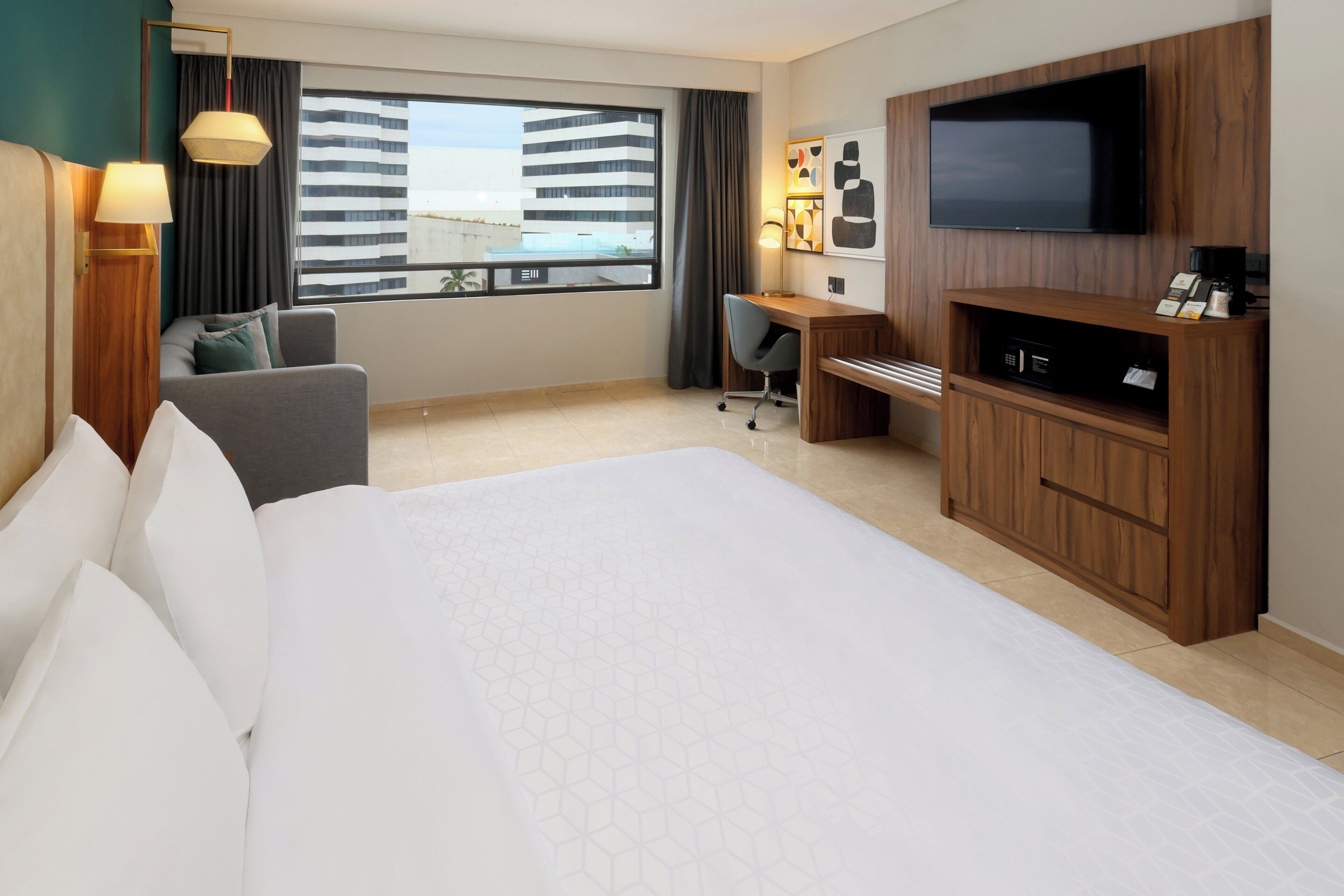 Ett hotellrum med en stor säng, ett skrivbord, en stol, en tv och utsikt över en byggnad genom fönstret.