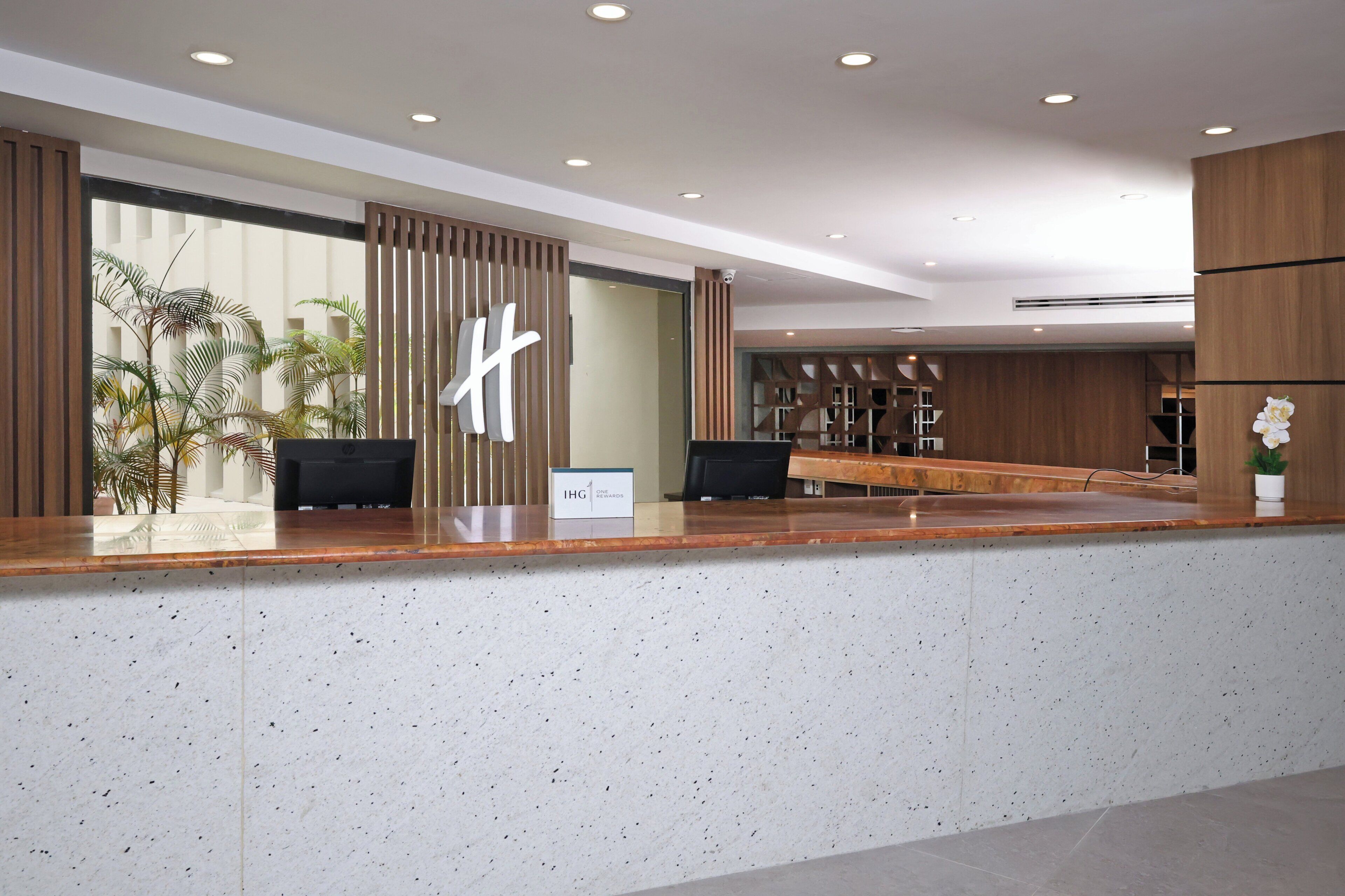 Un taulell de recepció modern amb un comptador de fusta, un senyal que diu "IHG" i vista de la zona del vestíbul amb plantes i una zona de seients.