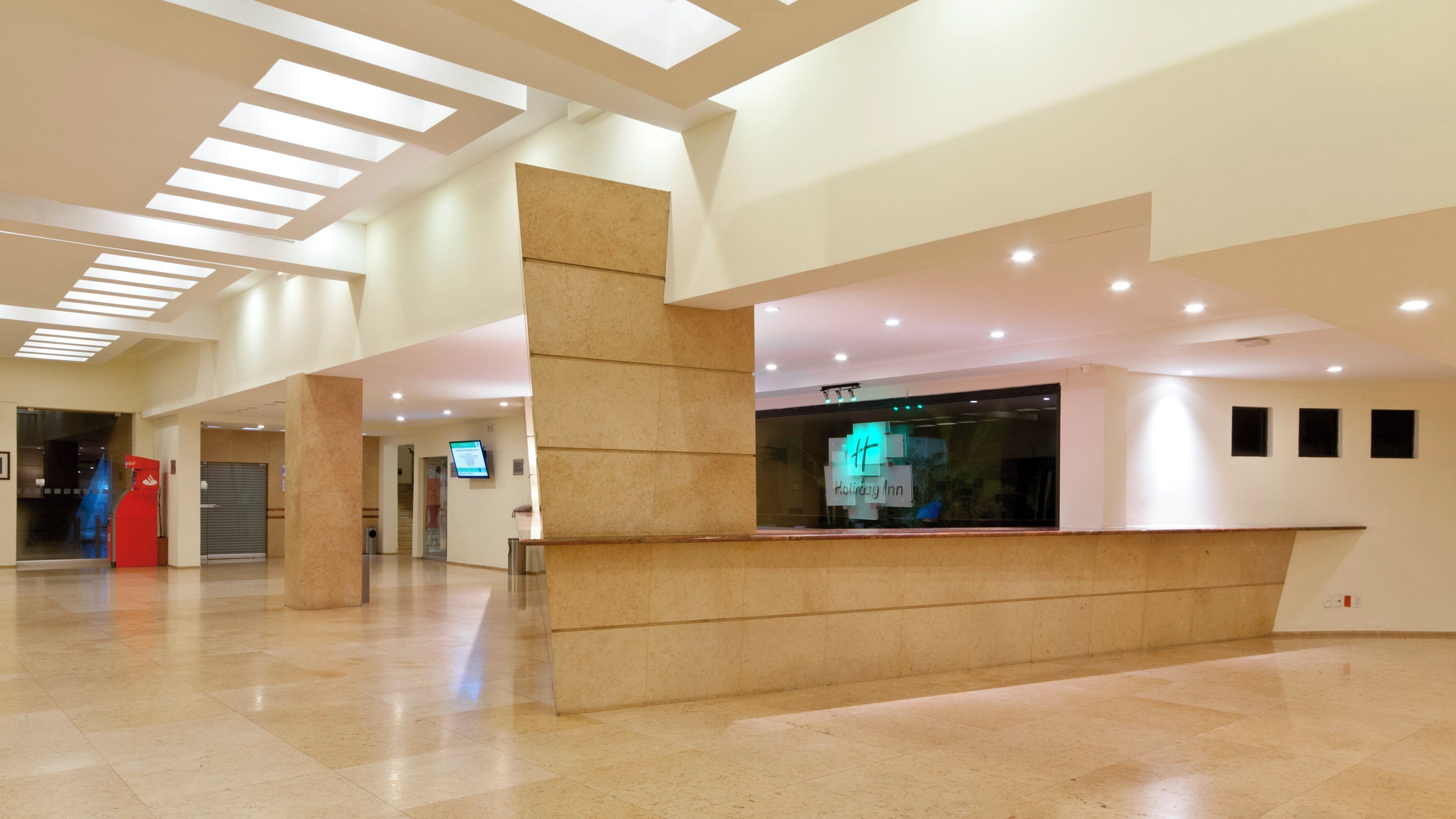 Un hall d’entrée spacieux comprenant un comptoir d’accueil, un panneau d’affichage numérique et un mur de verre sur lequel est apposée une enseigne indiquant un hôtel Holiday Inn.