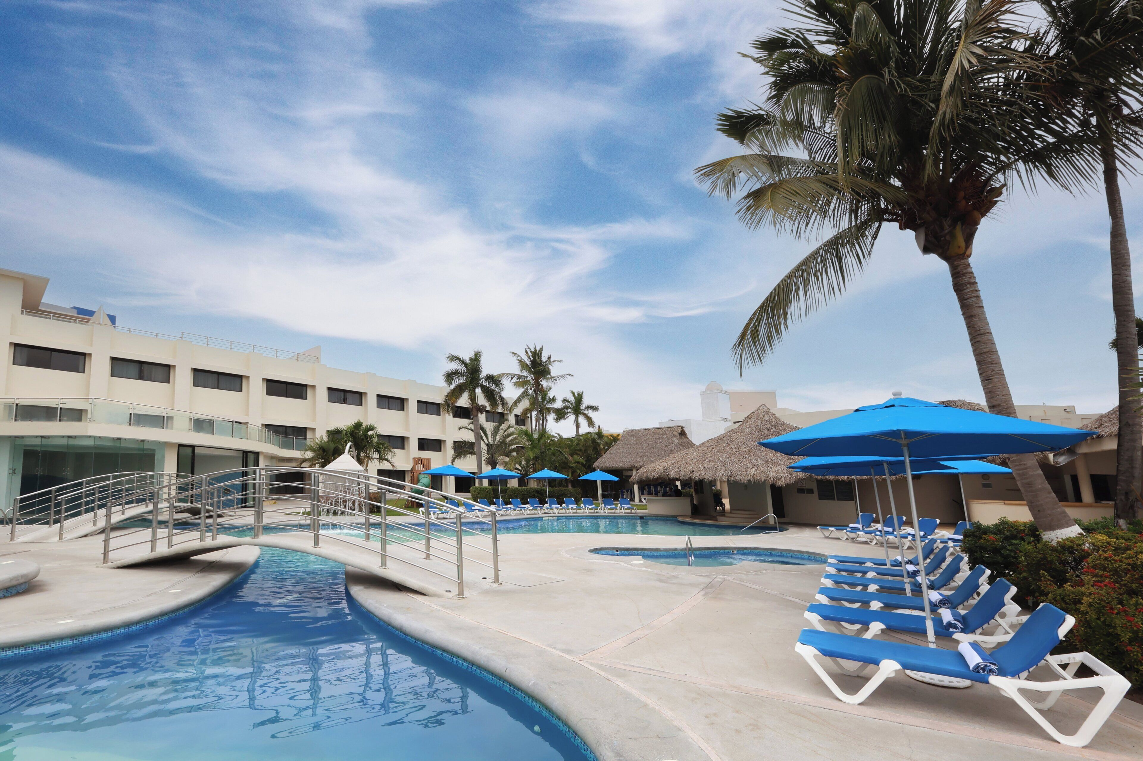 Zona de piscina en un resort con sillones de playa, sombrillas, palmeras y un edificio con varios balcones.