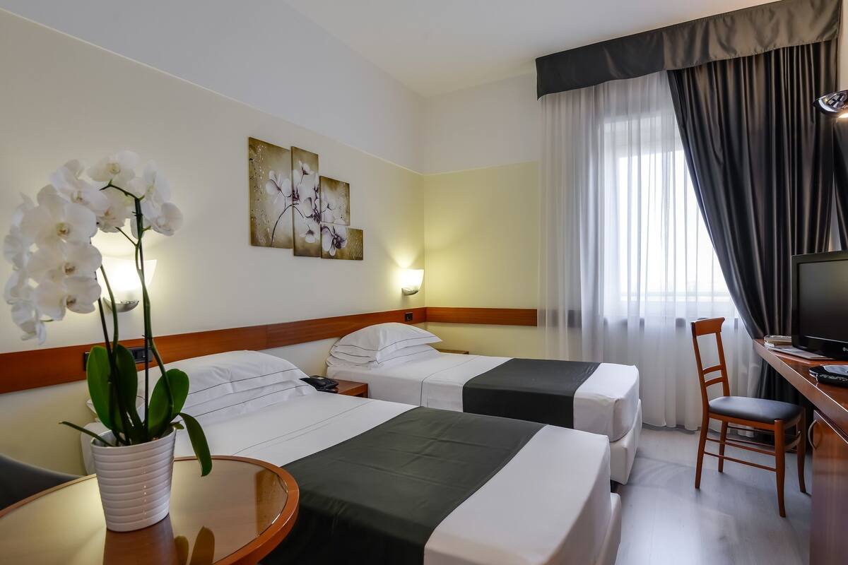 A hotel room with two beds, a desk, a chair, a TV, a lamp, and a potted orchid.