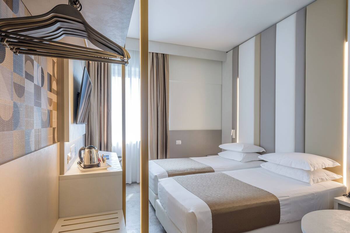 A hotel room with two beds, a desk with a kettle, a hanging coat rack, and a large window with curtains.
