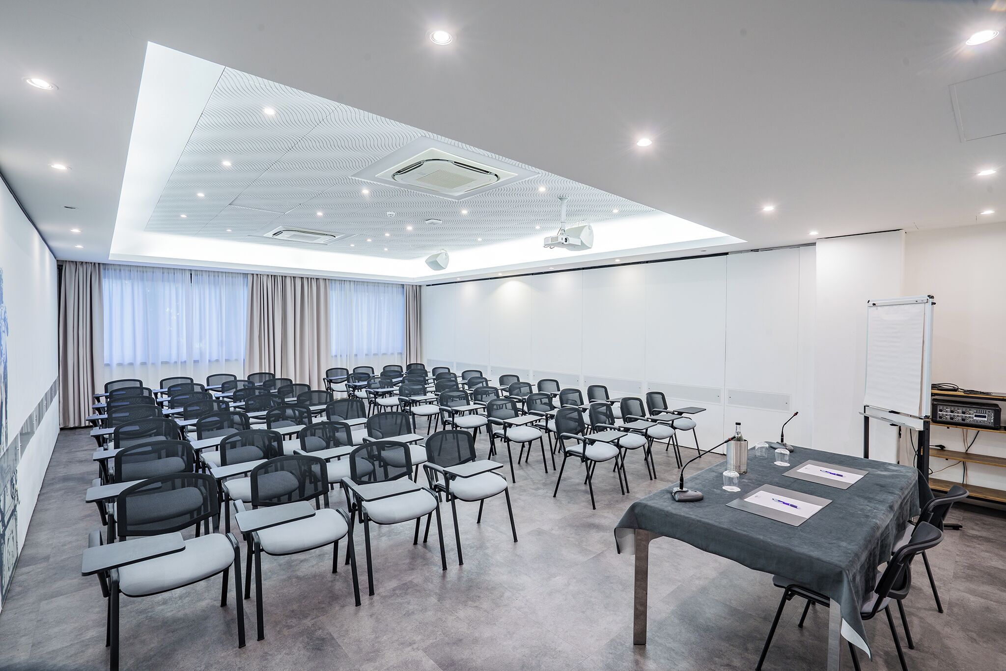 Ein Konferenzraum mit Reihen von Stühlen, einer Projektionsleinwand, einer Whiteboard-Tafel und einem Tisch mit Unterlagen und einem Mikrofon.