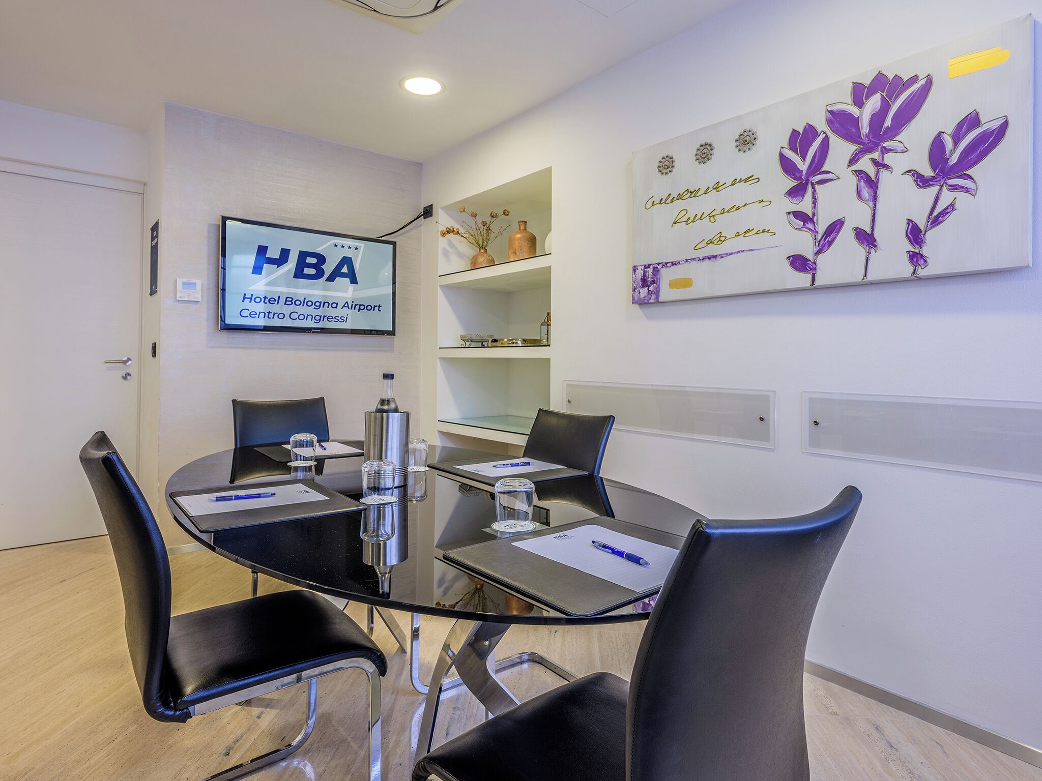 Un salón de conferencias con una mesa de vidrio, sillas negras, un televisor que muestra 'HBA Hotel Bologna Airport Centro Congressi' y una obra de arte en la pared con flores púrpuras.