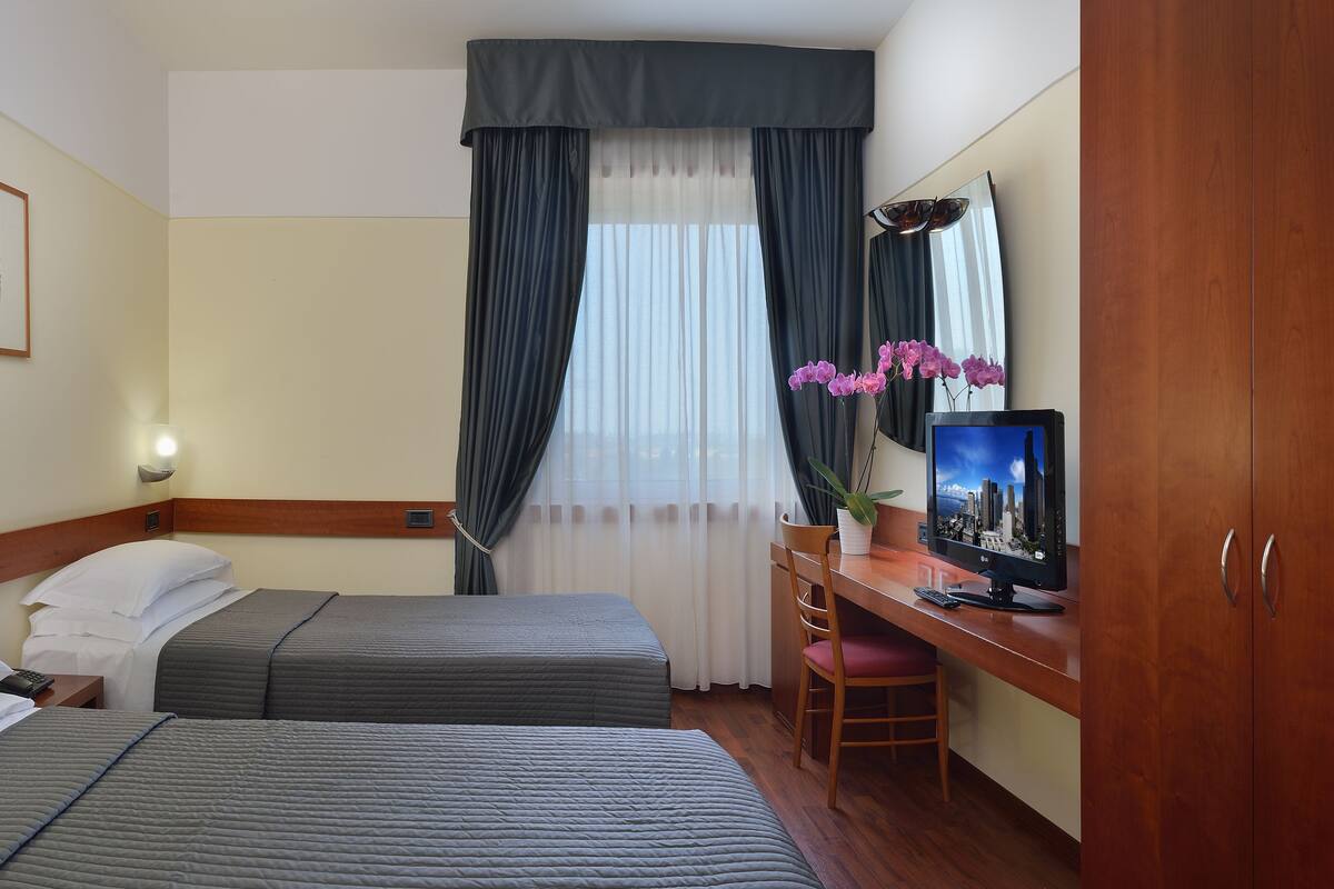 A hotel room with two beds, a desk with a computer, a mirror, and a window with curtains.