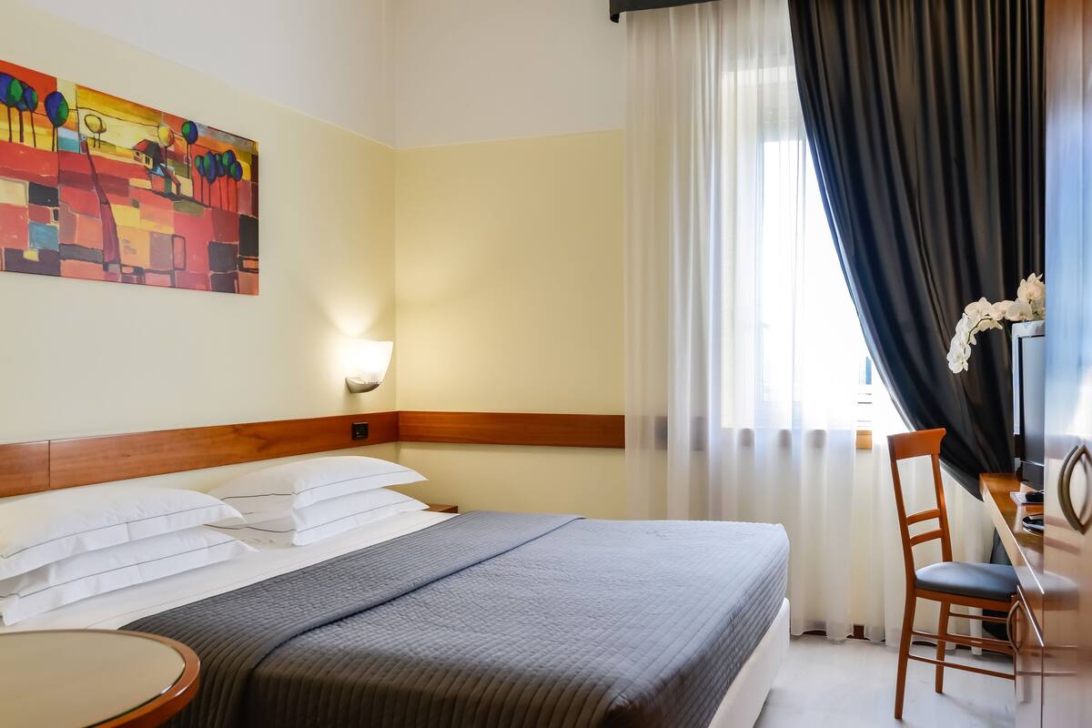 A hotel room with a bed, a desk, a chair, a painting on the wall, and a window with curtains.