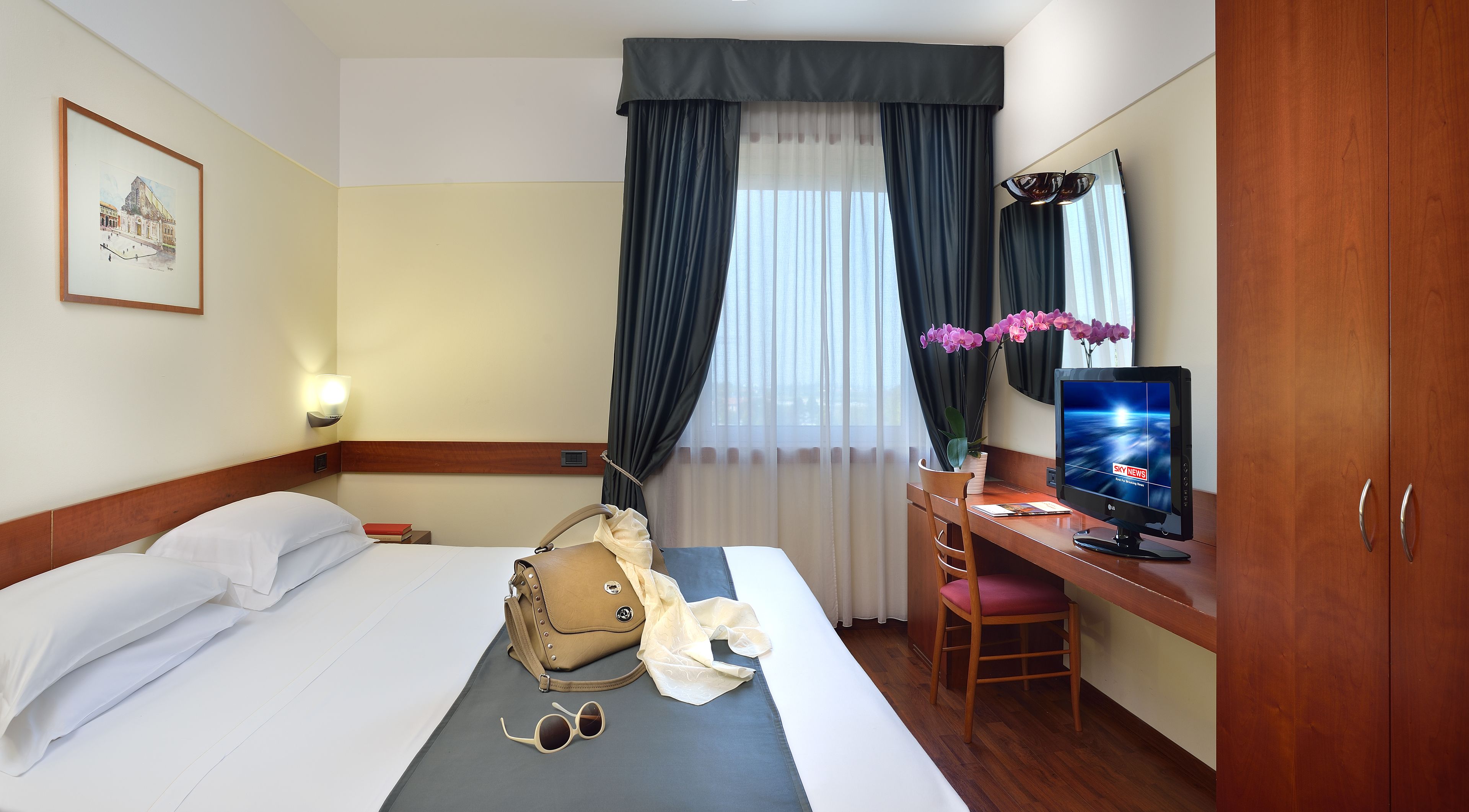 Una habitación de hotel con cama, escritorio, televisión y ventana con cortinas.
