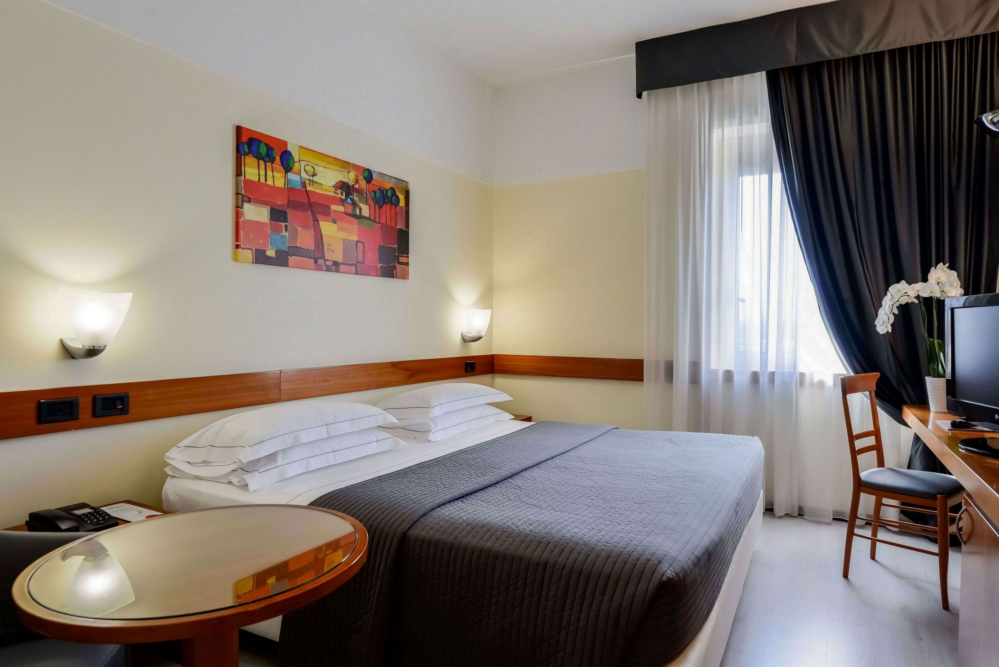 A hotel room with a bed, a desk with a chair, a small table, a TV, and a painting on the wall.