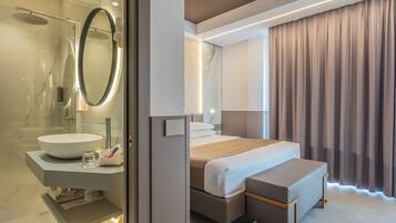 A modern hotel room with a glass-enclosed shower, a large round mirror, a white sink, a bed with a wooden bench, and a large window with curtains.