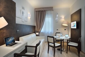 Una habitación de hotel moderna con un sofá blanco, un escritorio con una computadora portátil, una mesa de comedor con sillas y un televisor en la pared.