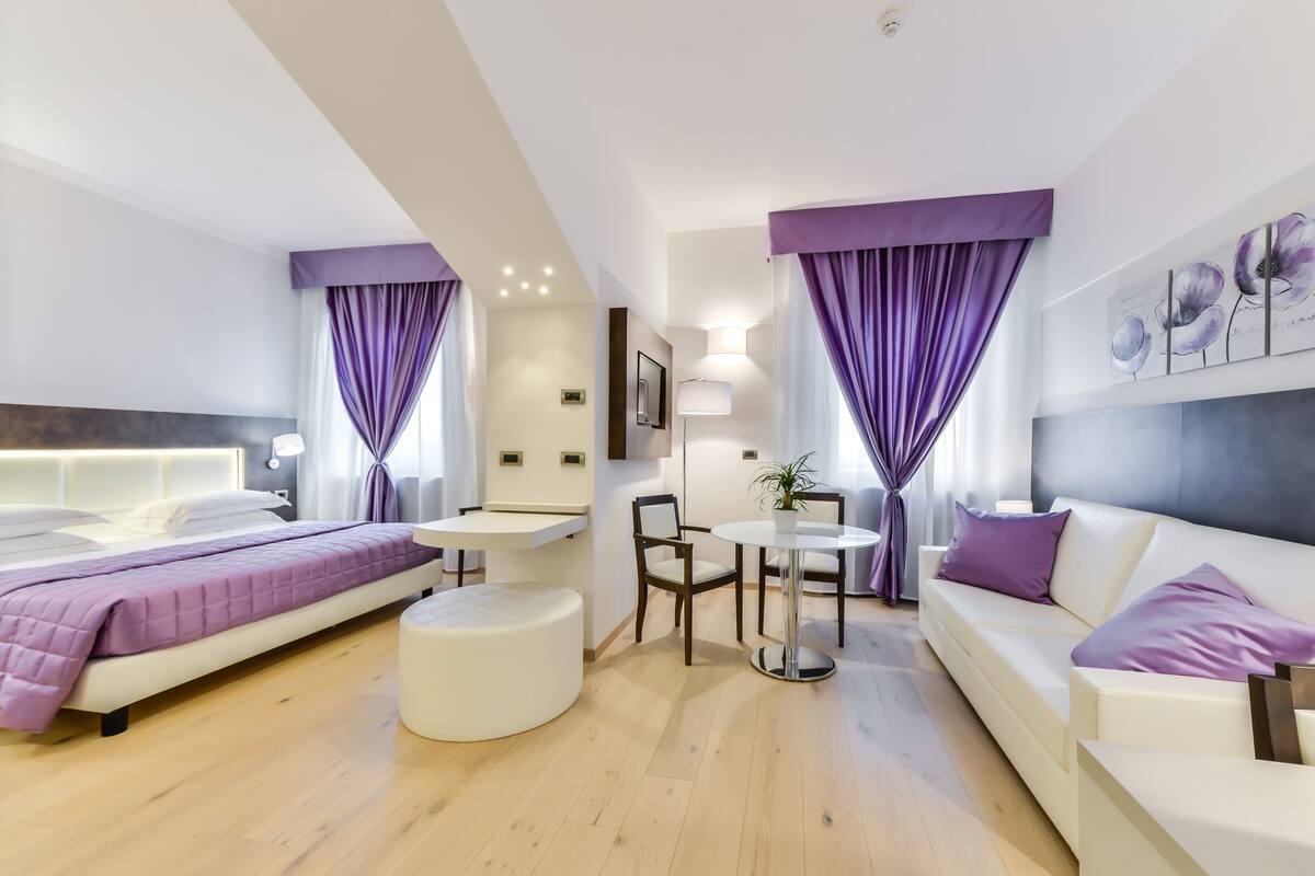 A modern hotel room with a bed, a sofa, a dining table, and a small seating area.