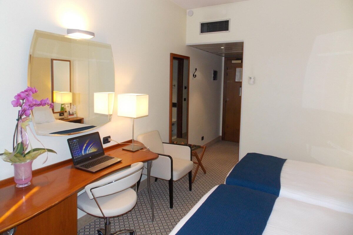 A hotel room with a desk, laptop, two bedside lamps, a bed, a chair, and a potted orchid.
