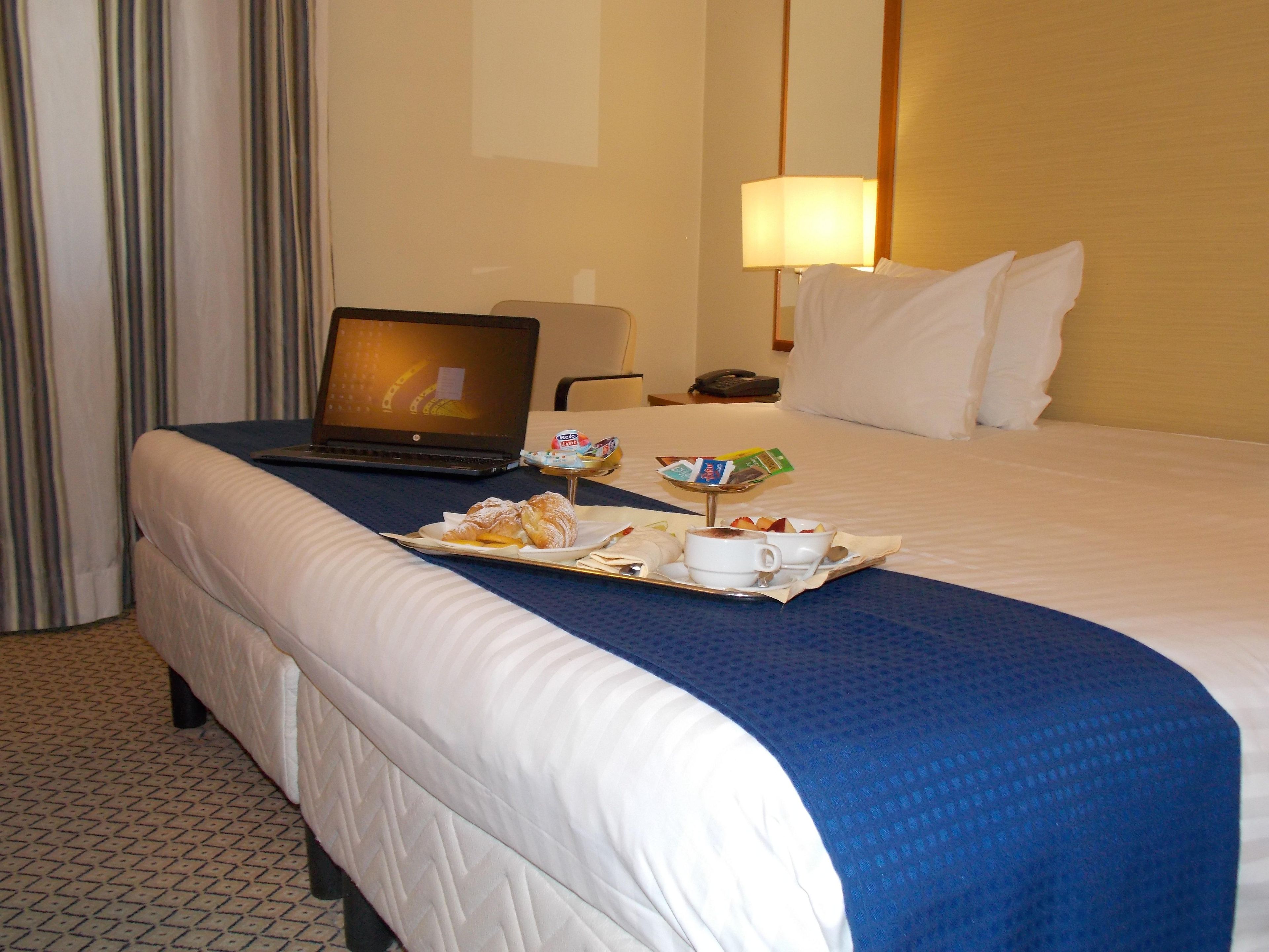 A hotel room with a bed, a laptop on the bed, a tray with food and a cup, and a lamp on the nightstand.