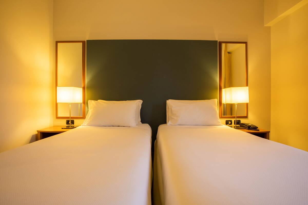 A hotel room with two single beds, a dark-colored headboard, bedside lamps, and a small table with a phone.