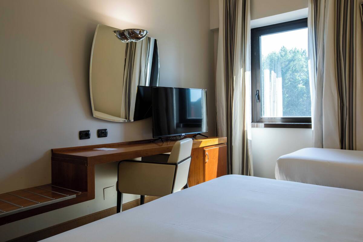 A hotel room with a bed, a desk with a chair, a television, and a window with curtains.