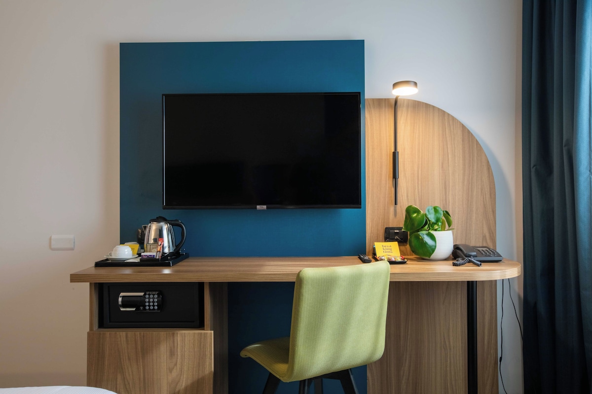 Une chambre d’hôtel avec un téléviseur à écran plat fixé à un mur bleu, un bureau en bois avec une chaise verte, une machine à café, une plante et une lampe.