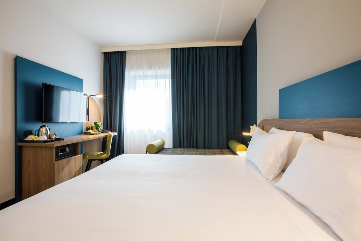 Une chambre d’hôtel avec un grand lit, un bureau avec une chaise, une télévision et une fenêtre avec des rideaux.