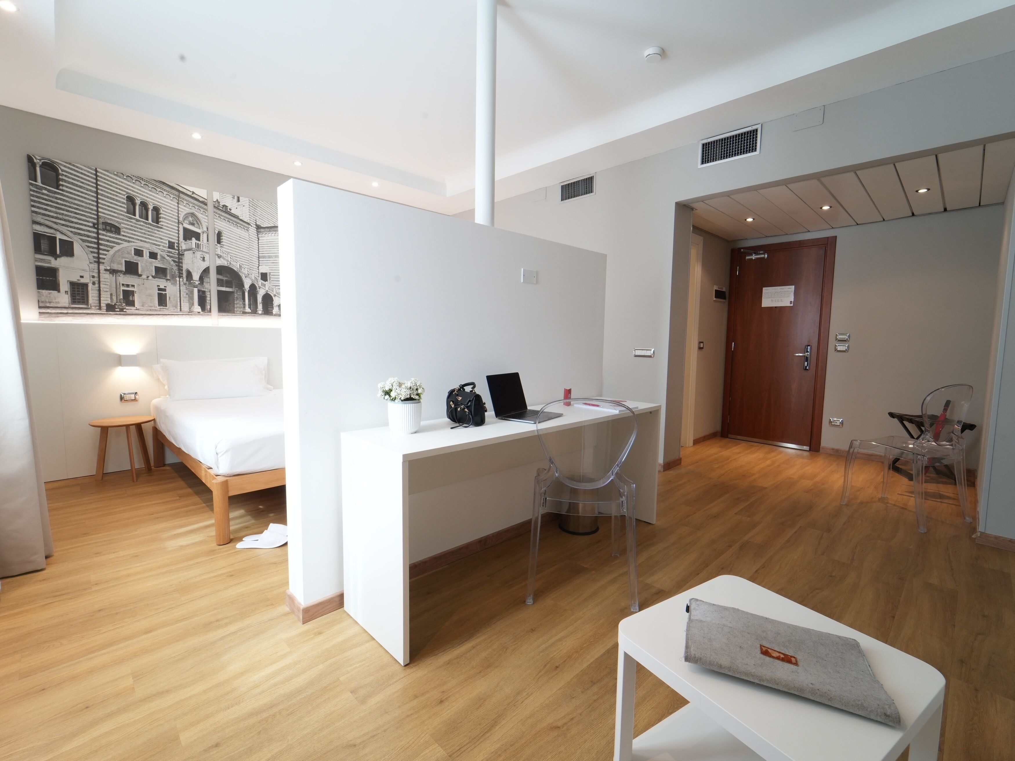 Ein modernes, helles Zimmer mit Holzboden, einem weißen Schreibtisch mit Laptop, einem Stuhl und einem Bett mit weißer Bettwäsche.