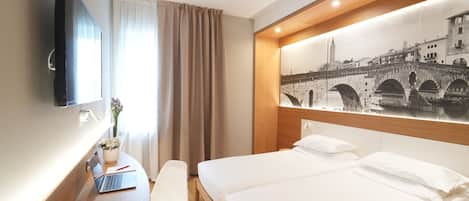 A hotel room with a bed, a desk with a laptop, a chair, a TV mounted on the wall, and a large window with curtains.