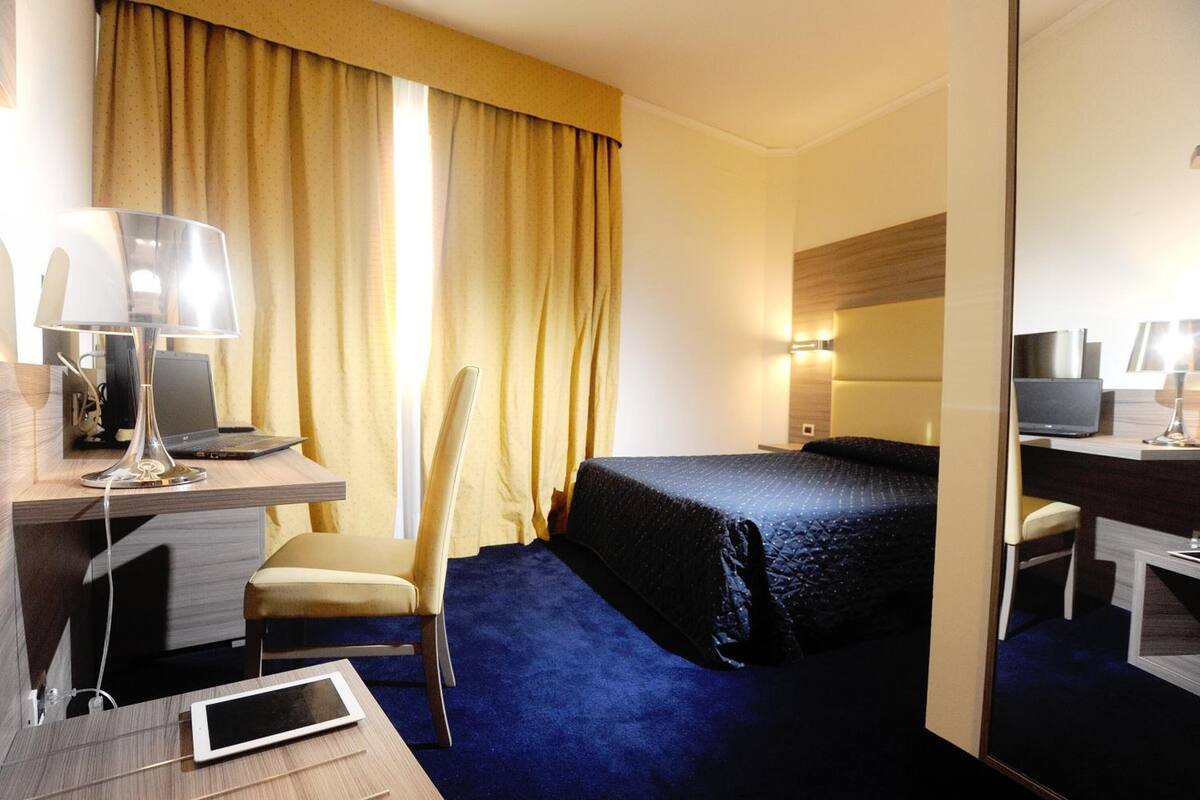 Habitación de hotel con cama, escritorio, silla, computadora portátil, tableta y cortinas.