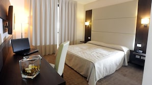 A hotel room with a bed, a desk with a laptop, a glass of water, a chair, and a nightstand with a phone. - Palace Inn (Fiano Romano)