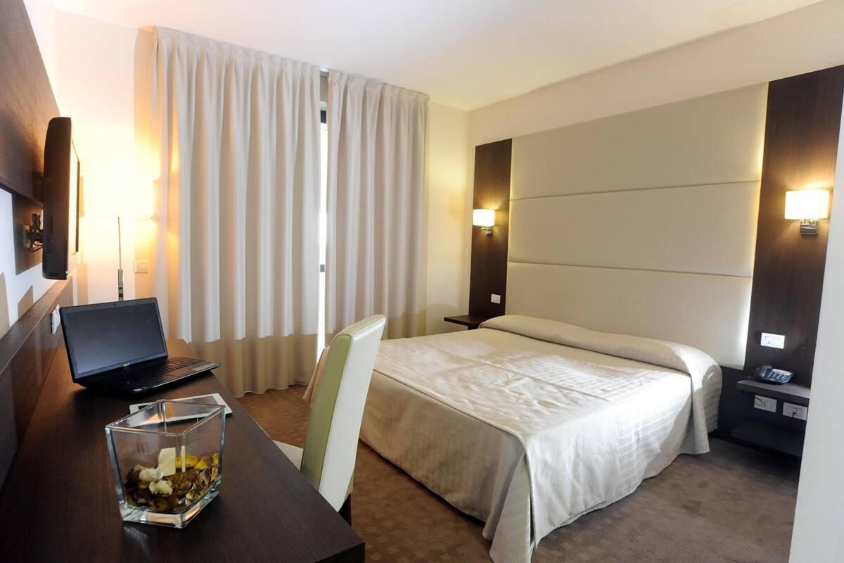 Habitación de hotel con una cama, un escritorio con una computadora portátil, un vaso de agua, una silla y una mesita de noche con un teléfono.