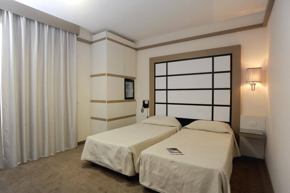 Habitación de hotel con dos camas, un ventanal con persianas, una mesita de noche con lámpara y un teléfono en la pared.