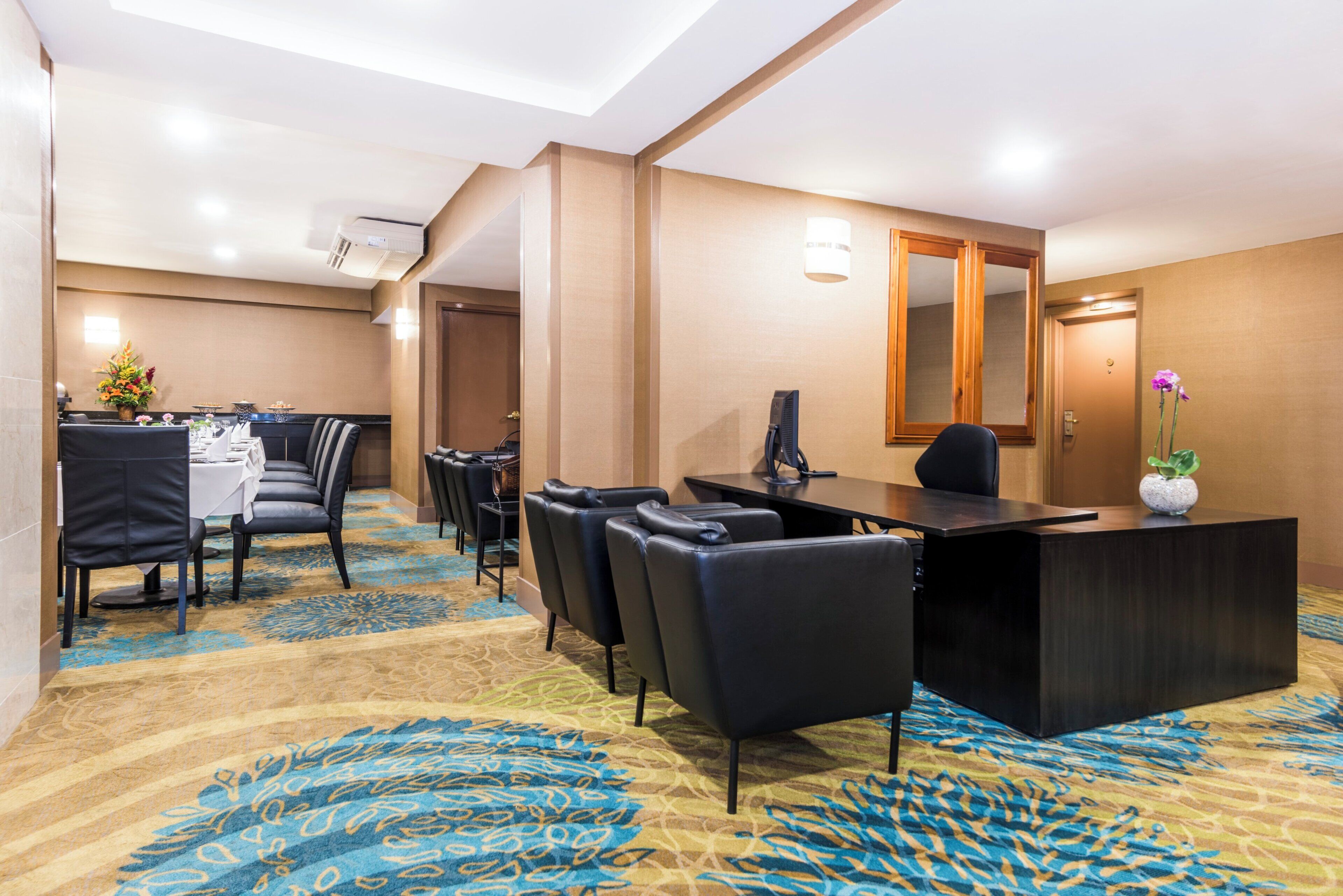 A hotel conference room with a reception desk, several chairs, a table with a white tablecloth, and a patterned carpet.