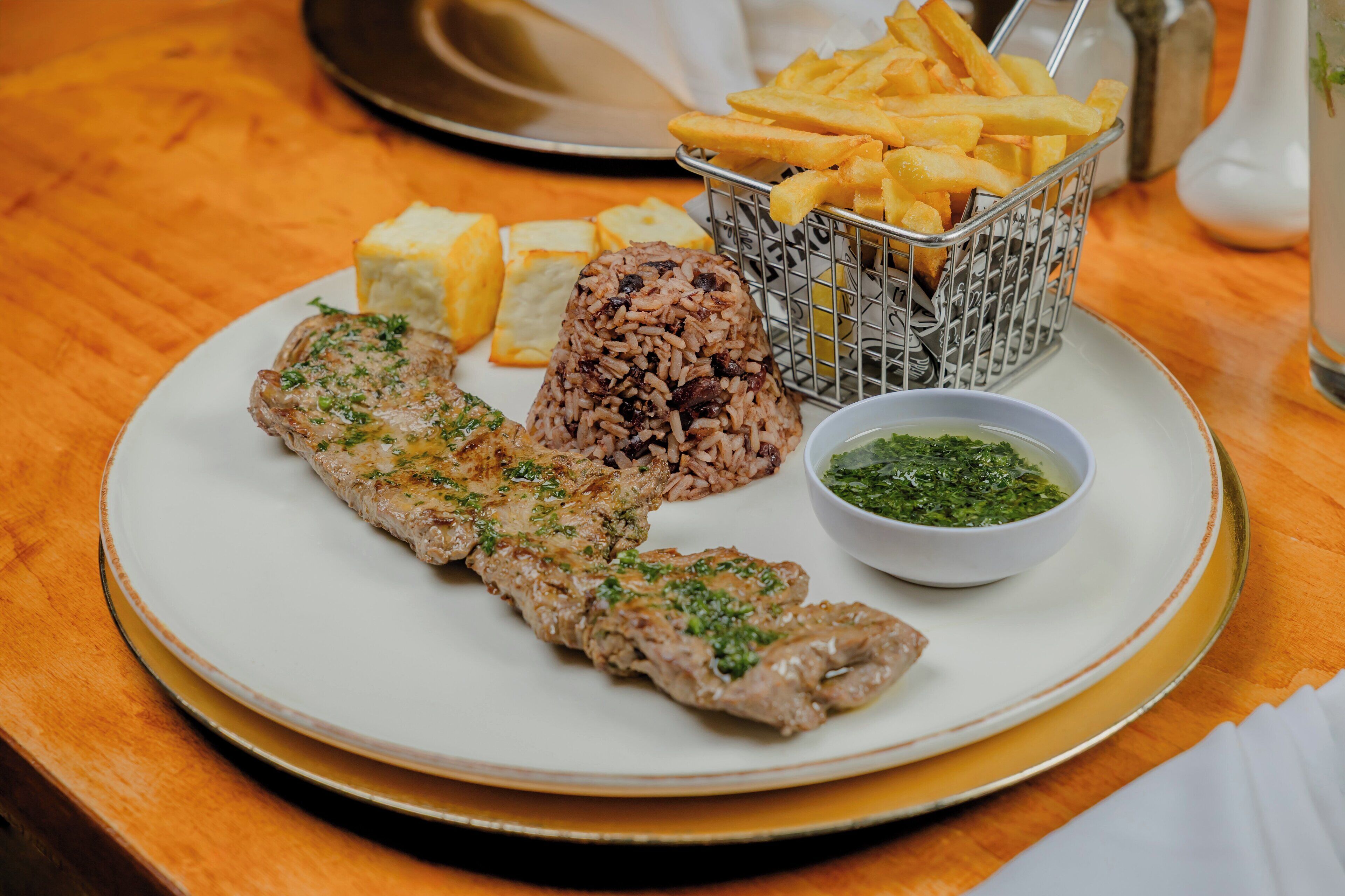 A plate with a meat dish, rice, fries, and a green sauce.