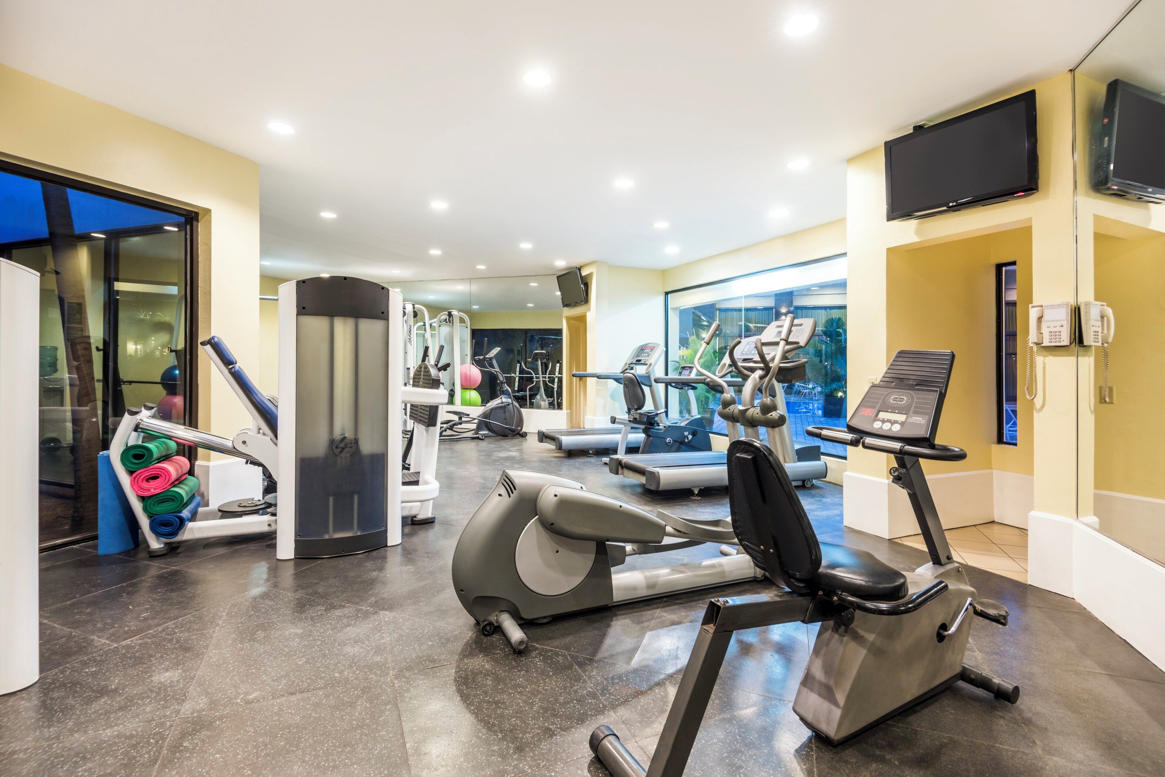 Un gimnasio con equipo de ejercicio diverso, cinta de correr y pared con espejos.